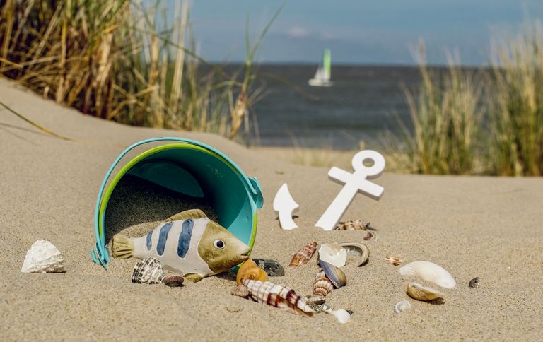 Playful beach scene with a toy bucket, seashells, and a distant sailboat on a sunny day.